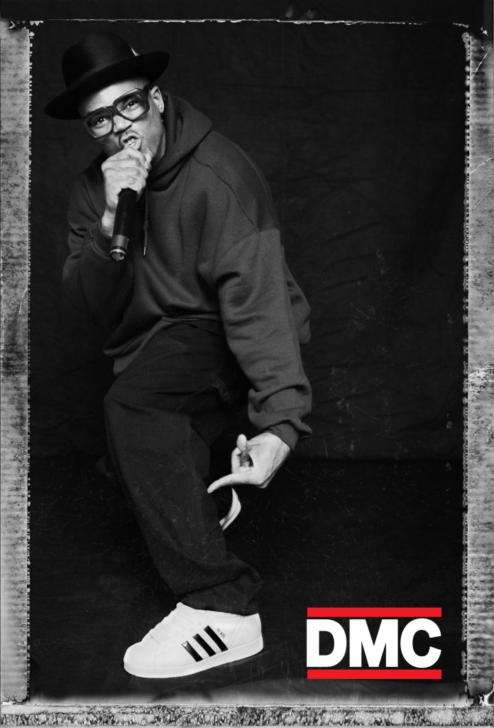 Black and white photo of Darryl "DMC" McDaniels of Run-DMC, holding a microphone. The letters "DMC" are written in white, and outlined in red, in the bottom righthand corner.
